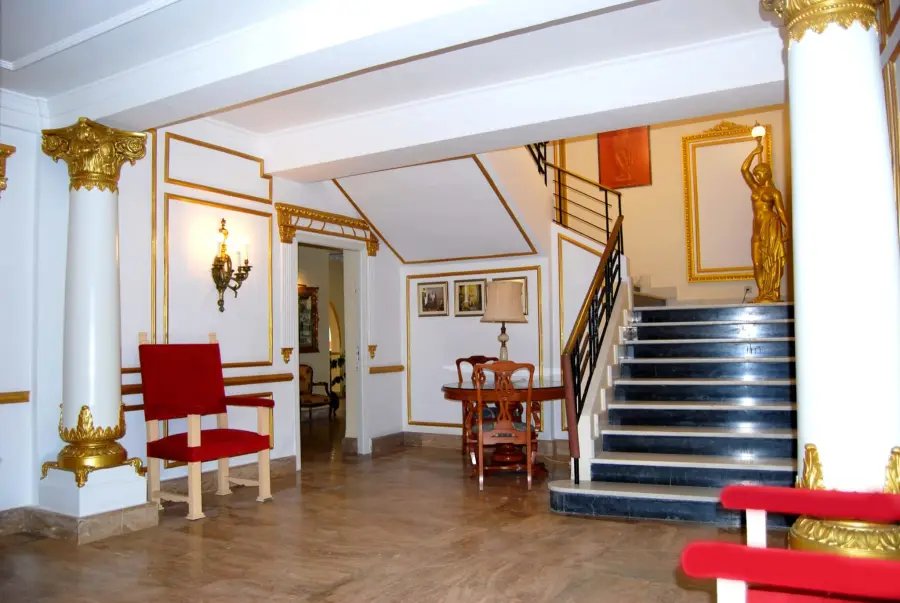 Hall con columnas blancas y doradas, sillón rojo y escalera